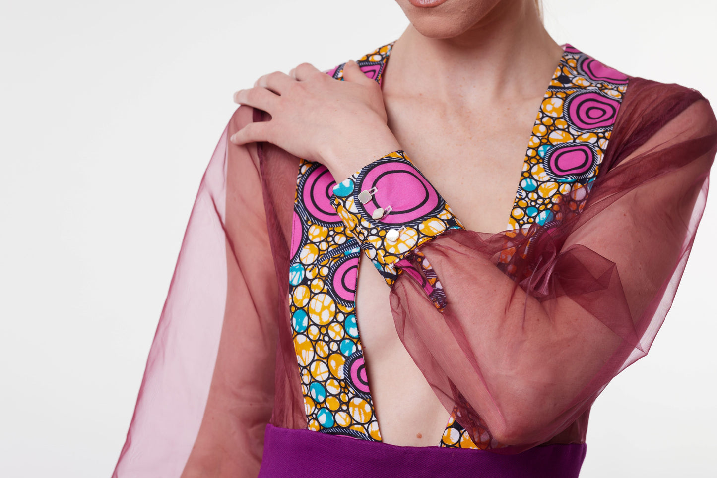 combinaison bordeaux avec un col plongeant orné de motifs wax roses, jaunes et bleus, et des manches transparentes en organza bordeaux, posant avec les bras croisés.