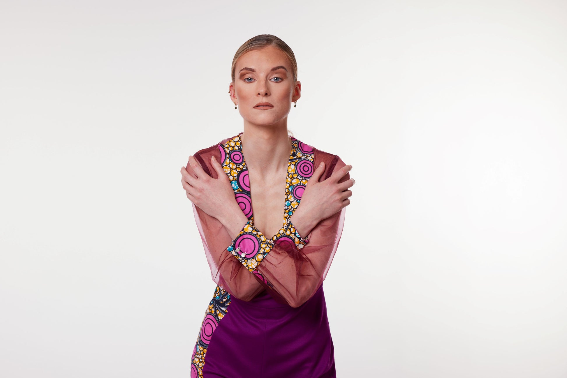 Mannequin posant bras croisés, portant un haut en organza de soie transparente bordeaux avec un col plongeant orné de motifs wax aux cercles roses, jaunes et bleus, devant un fond neutre.